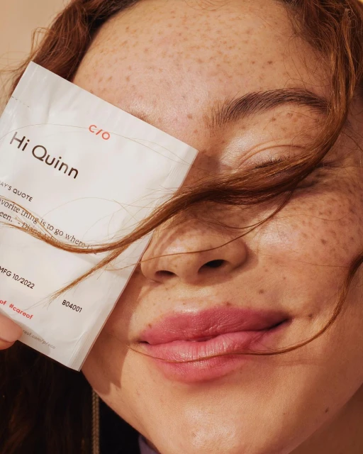 Close-up shot of a red-haired, freckled woman holding a white product packet with the brand name 'Hi Quinn' partially obscuring her face.