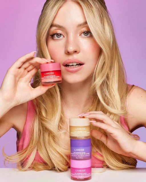 Sydney Sweeney poses with a stack of Laneige lip sleeping masks. Pink and purple background. Beauty and skincare concept.