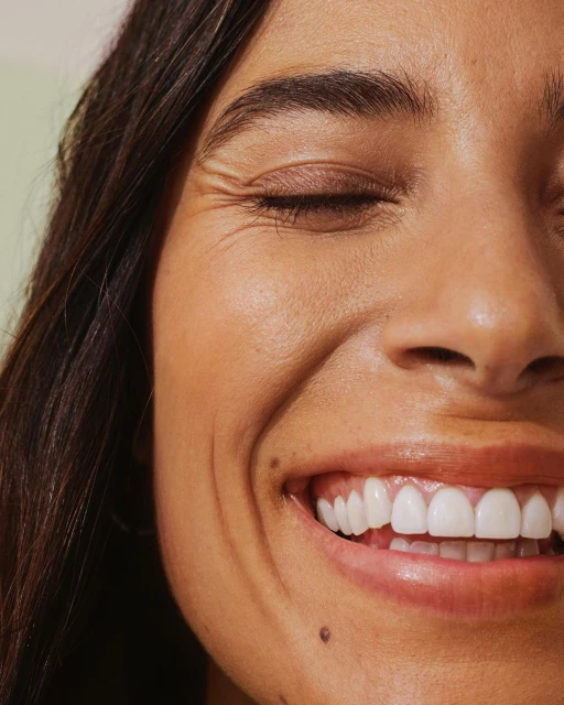 Close-up of a smiling woman with dark hair and light skin showcasing perfect white teeth against a soft green background.