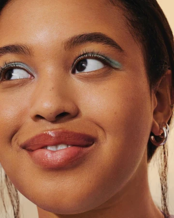 Close-up portrait of a Black woman with glossy lips, green eyeshadow, and silver hoop earrings against a warm background.
