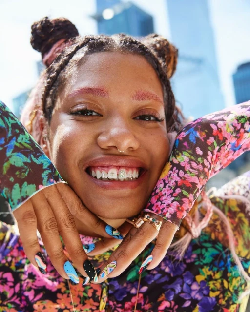 Close-up portrait of a smiling woman with styled hair, floral top, and decorative braces posing in an urban outdoor setting.