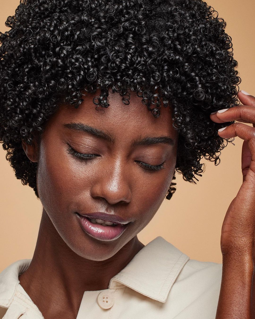 Close-up of a Black woman with curly hair, tan skin, and light-colored top, set against a neutral backdrop in a beauty-style portrait.