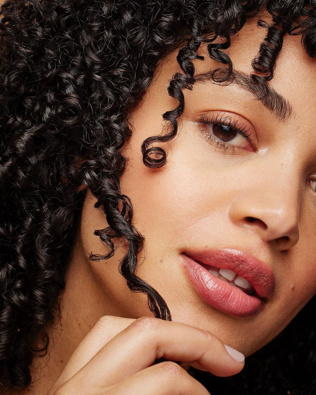 Close-up portrait of a young adult female with curly black hair, light skin, and pink lipstick, posing indoors under bright lighting.