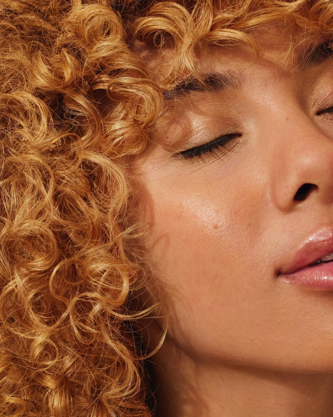 Close-up beauty portrait of a woman with curly blonde hair and closed eyes in soft natural light.