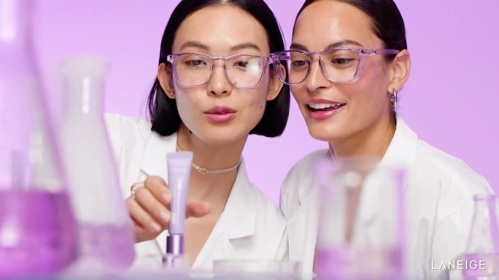 Two women in lab coats examine a beauty product against a light purple background, surrounded by lab glassware. Beauty advertising.