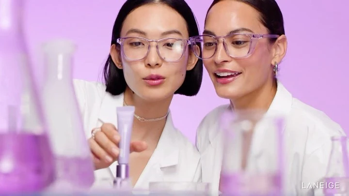 Two women in lab coats examine a beauty product against a light purple background, surrounded by lab glassware. Beauty advertising.