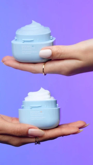 Two light blue cosmetic cream containers, each held by different hands, stand out against a vibrant blue and purple gradient background.