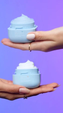 Two light blue cosmetic cream containers, each held by different hands, stand out against a vibrant blue and purple gradient background.