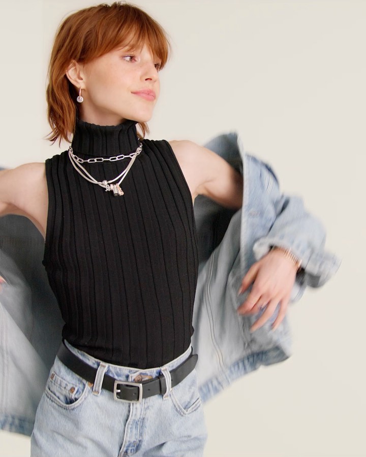 A woman with red hair wears a black turtleneck and light blue jeans, accessorized with layered silver necklaces and a denim jacket against a neutral backdrop.