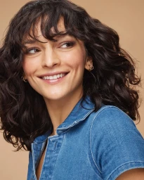 Portrait of a smiling woman with dark curly hair wearing a blue denim shirt against a warm neutral background