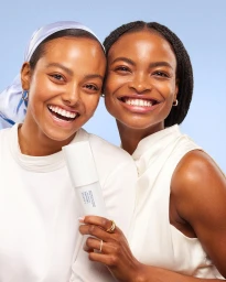 Two smiling Black women holding a skincare bottle against a solid blue background, photographed by Mckenzie Thompson.