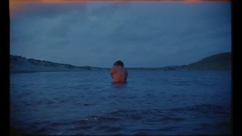 Person stands in calm blue ocean water at dusk. Hilly landscapes form a horizon under a cloudy blue sky, captured at water-level.