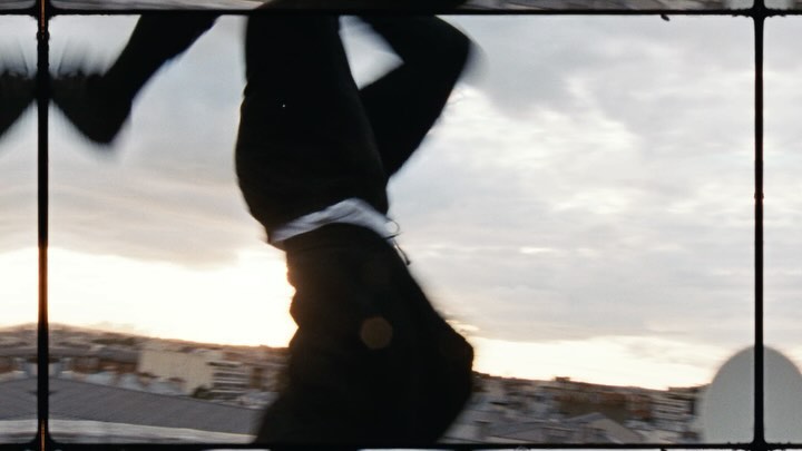 A person in black clothing is upside down against a cloudy sky and city buildings, framed by a window-like structure, creating an abstract urban image.