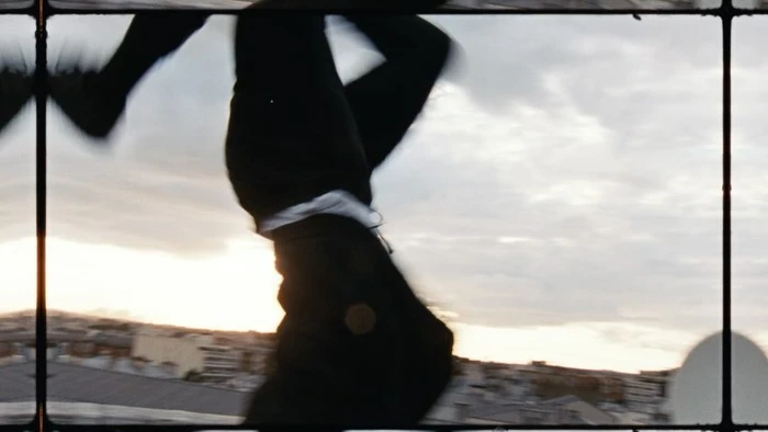 Inverted film frame showing a person suspended upside down against a cloudy sky above urban rooftops, directed by Mees Peijnenburg.