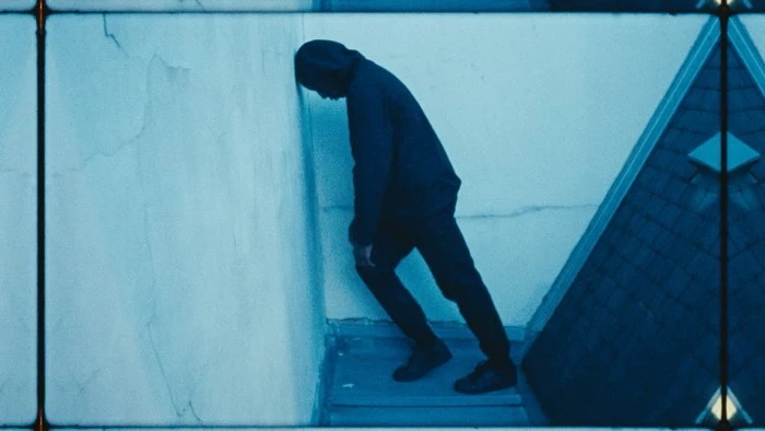 A person in a dark hooded jacket leaning head against a wall on a rooftop under a monochromatic blue color grade.