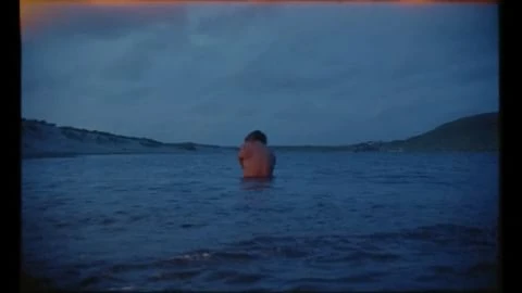 A person submerged up to the chest in a calm dark body of water at dusk with rolling hills along the shoreline.