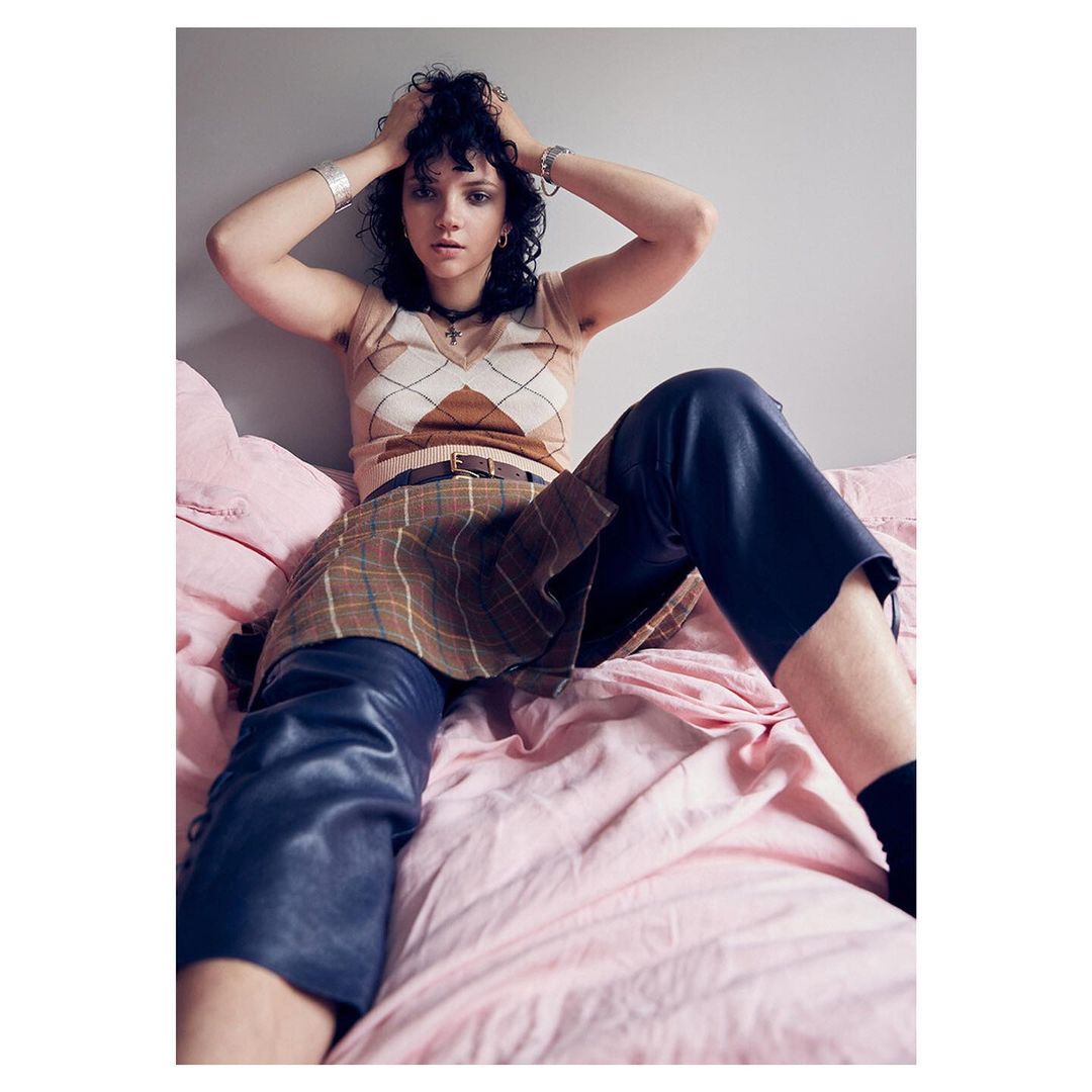 Young adult woman in a patterned sweater and skirt poses on a pink bed in a studio-style shot with a neutral wall backdrop.