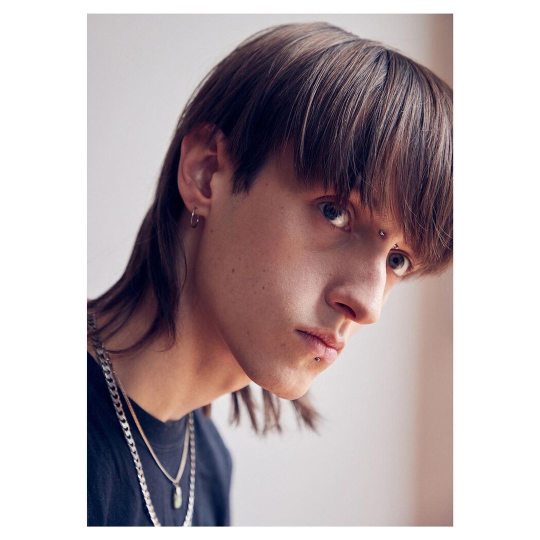 Close-up portrait of a young adult male with dark hair, light eyes, pierced eyebrow and lip, wearing a dark shirt and layered necklaces.