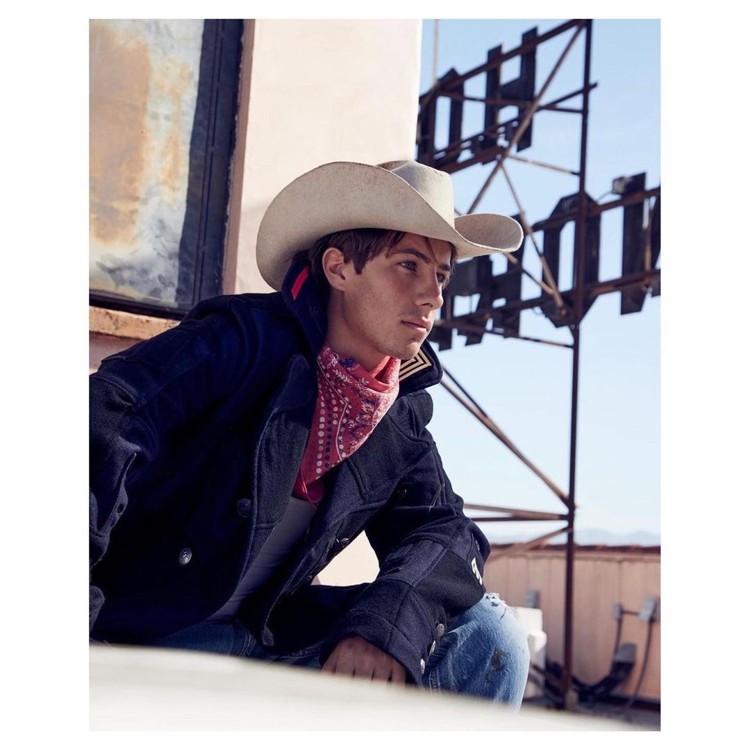 Young adult in cowboy hat and jacket poses near rusted metal structure in an outdoor setting with clear blue sky in the background.