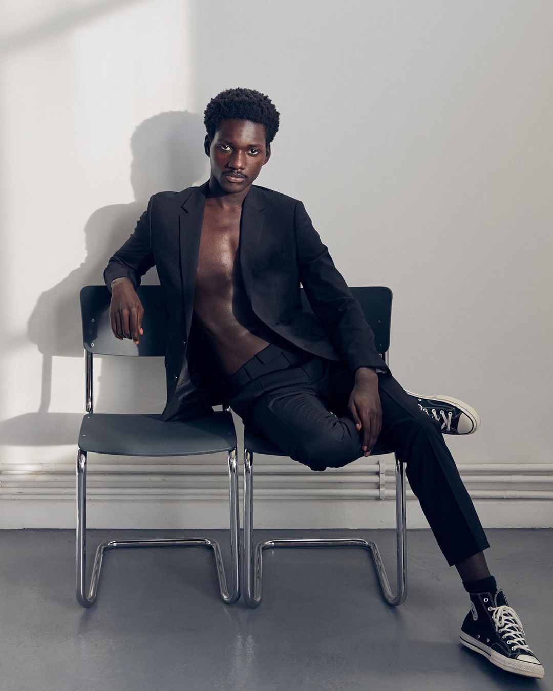 Adult male model poses indoors on gray chair wearing a black suit with no shirt. He has black sneakers. White wall background.