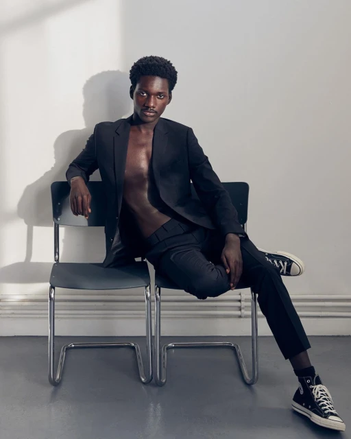 Adult male model poses indoors on gray chair wearing a black suit with no shirt. He has black sneakers. White wall background.