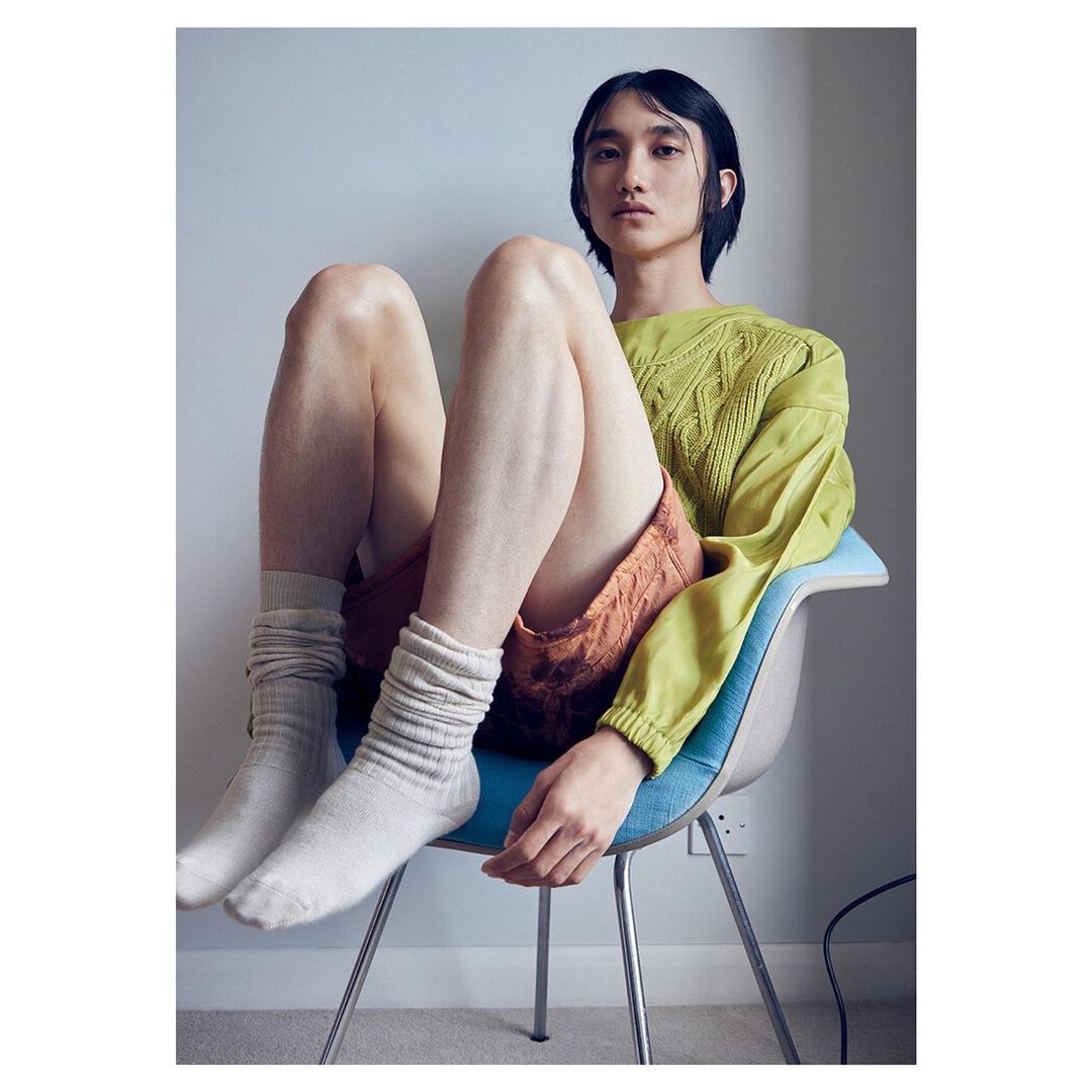 Portrait of an adult male sitting in a blue chair wearing socks, a light green sweater, and rust-colored shorts indoors.