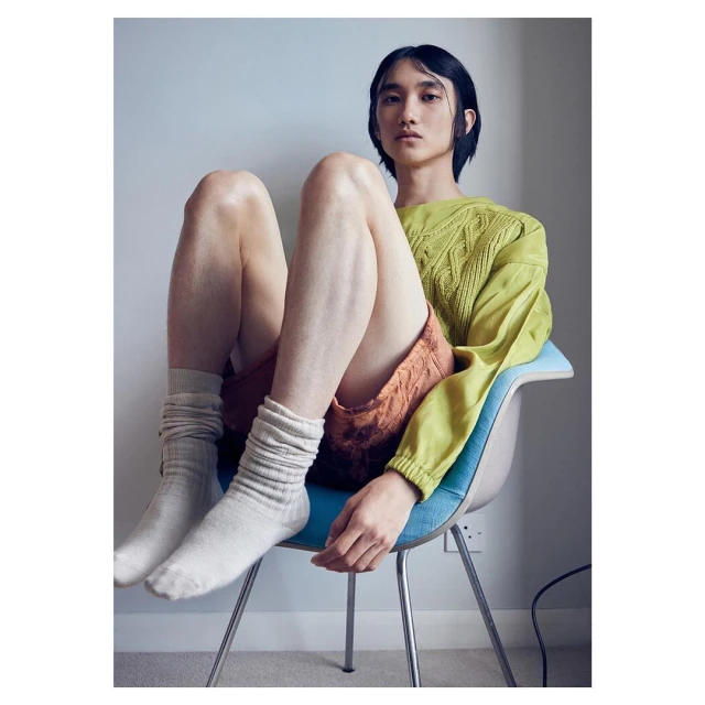 Portrait of an adult male sitting in a blue chair wearing socks, a light green sweater, and rust-colored shorts indoors.