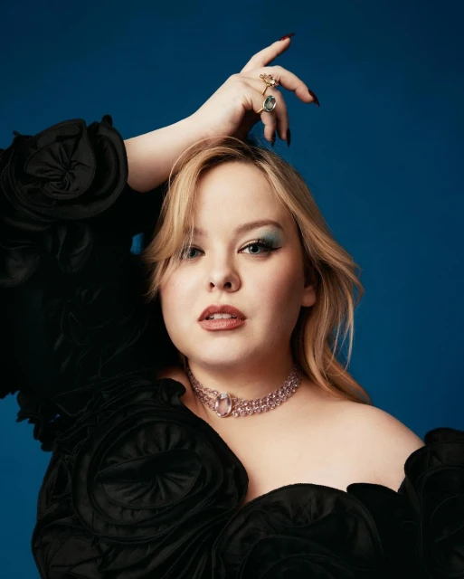 Portrait of a blonde woman with blue eyeshadow and pink jewelry against a solid blue background, wearing a black dress.