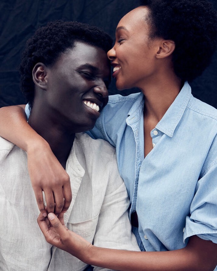 Intimate portrait of a smiling African American couple in light-colored denim shirts embracing against a dark backdrop in a close-up shot.