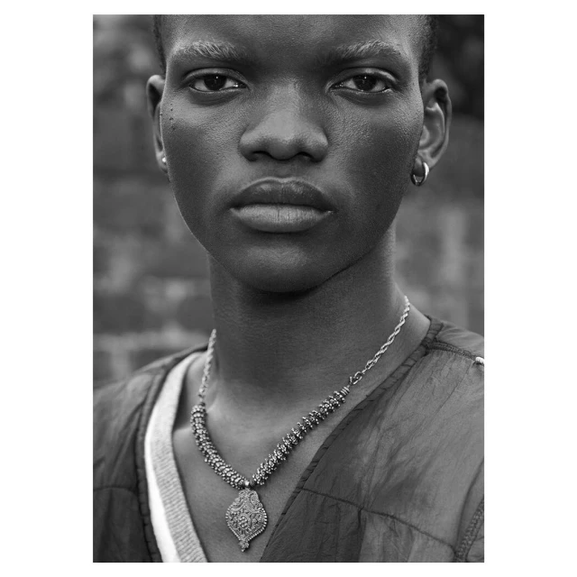 Close-up black and white portrait of a young adult Black male with a serious facial expression wearing a necklace against a brick wall.