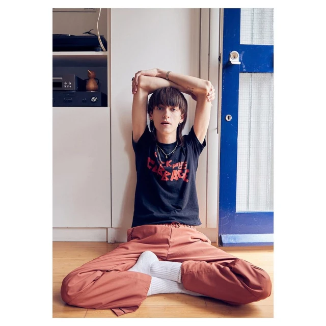 A young adult sits in an indoor studio wearing a graphic tee and red-orange pants. They are leaning back against a wall.