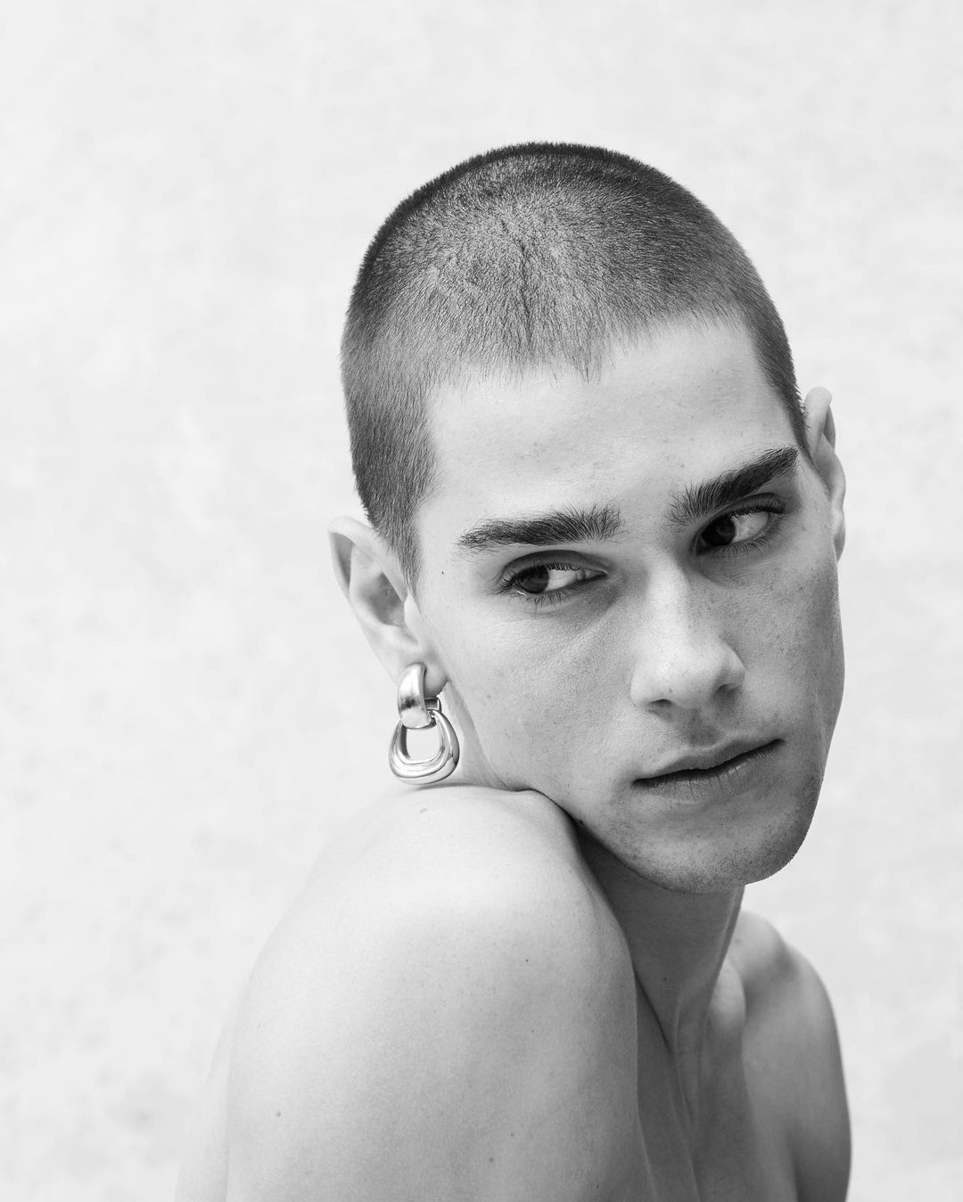 Monochrome portrait of a young adult male with a shaved head, posing with bare shoulders and wearing a silver earring against a plain backdrop.