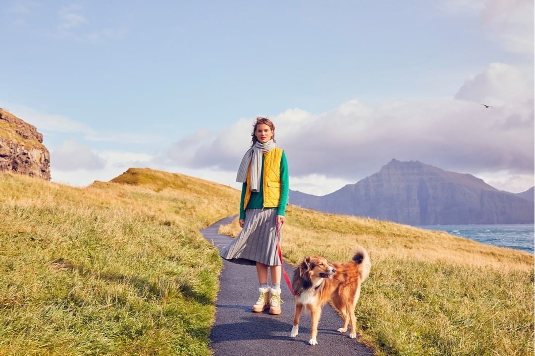 A woman walks her dog on a path through a grassy field with mountains in the background on a sunny day.