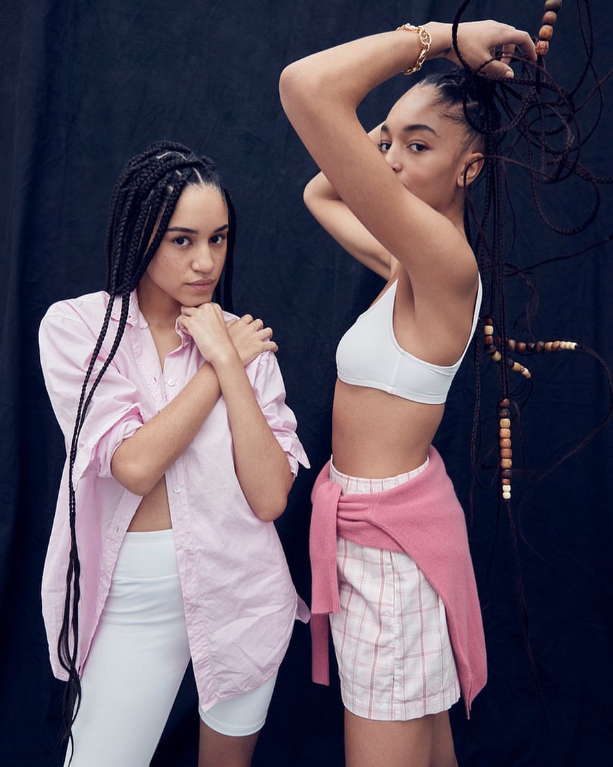 Two women with box braids pose against a dark backdrop, wearing casual tops and shorts, one with a sweater tied around her waist.