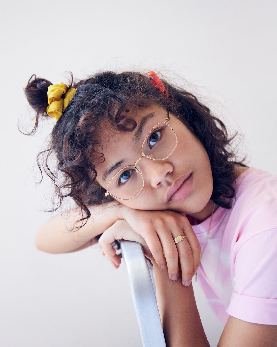 Portrait of a young female with curly dark hair and glasses, wearing a pink T-shirt, resting her arms on the back of a silver chair.
