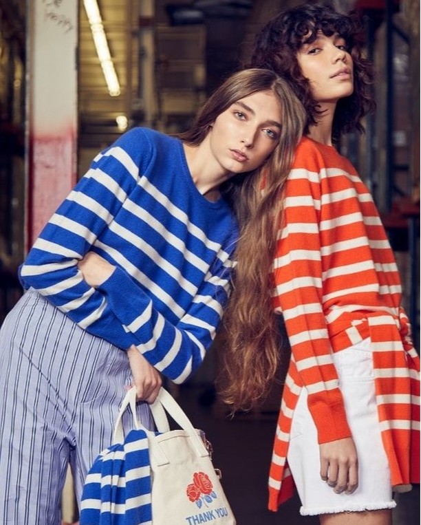 Two fashion models pose indoors wearing blue and orange striped sweaters with light-blue striped pants and a white tote bag.