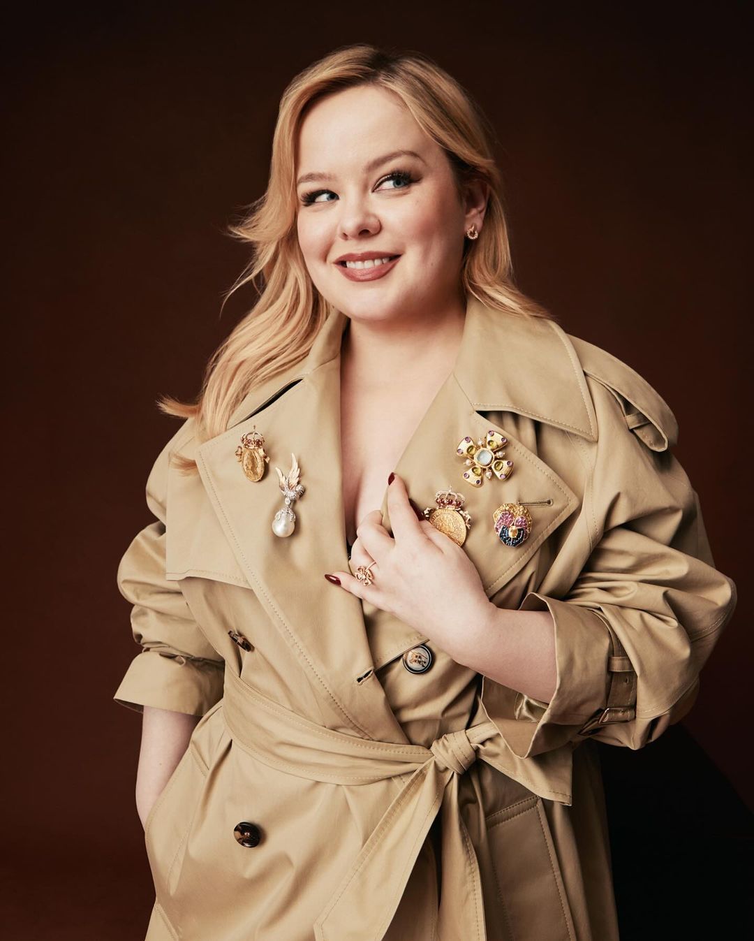 Portrait of a blond adult female in a beige trench coat adorned with vintage brooches, posing against a brown background in a photo studio.