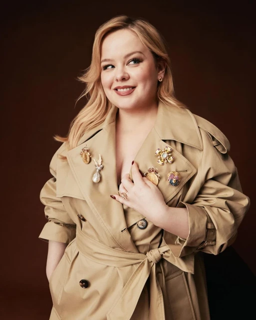 Portrait of a blond adult female in a beige trench coat adorned with vintage brooches, posing against a brown background in a photo studio.
