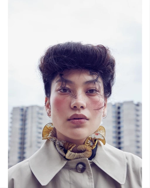 Close-up fashion portrait of an Asian woman with dark hair wearing a trench coat and ruffle collar against an urban background.