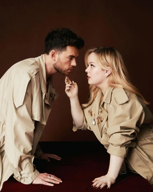 A close-up of a fashionable couple, an adult male and female, wearing tan trench coats looking at each other in a studio setting.