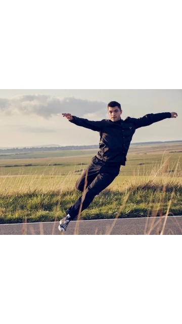 Athletic man in black tracksuit performs an acrobatic jump on a rural road, showcasing dynamic movement against a natural backdrop.