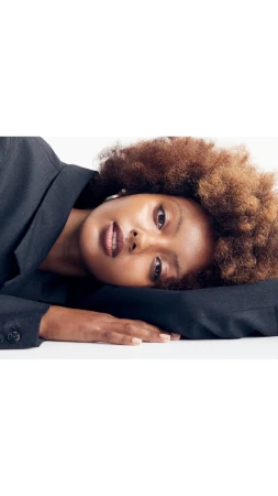 Portrait of a Black woman with voluminous curly hair wearing a dark blazer, lying down against a white background.