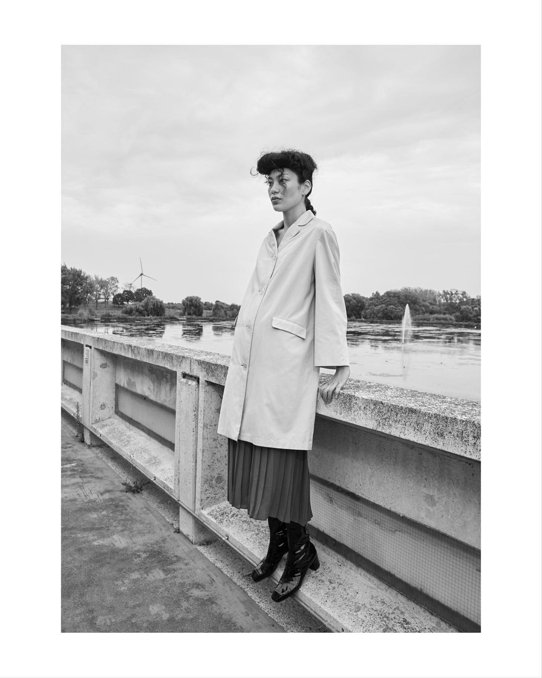 Monochrome portrait of a woman in an oversized coat and skirt standing on a bridge with a lake and wind turbines in the background.