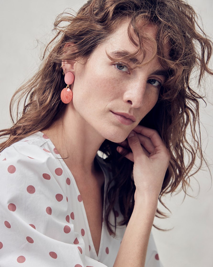 Close-up of a woman with wavy brunette hair wearing pink earrings and a white blouse with pink polka dots, looking at the camera.