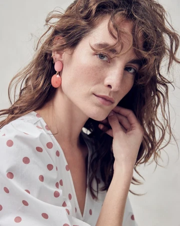 Close-up of a woman with wavy brunette hair wearing pink earrings and a white blouse with pink polka dots, looking at the camera.