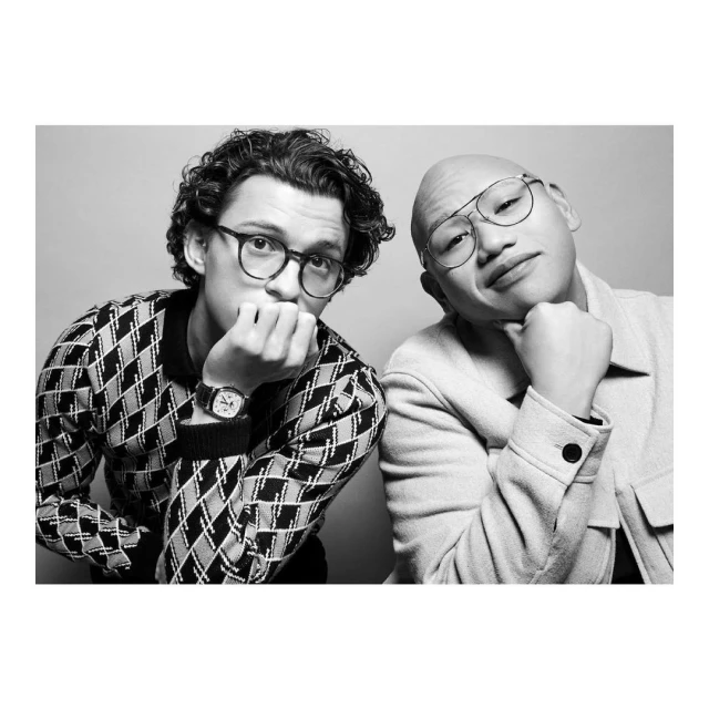 Black and white portrait of two men indoors. One with dark curly hair and glasses, the other with a shaved head and wire-frame glasses.
