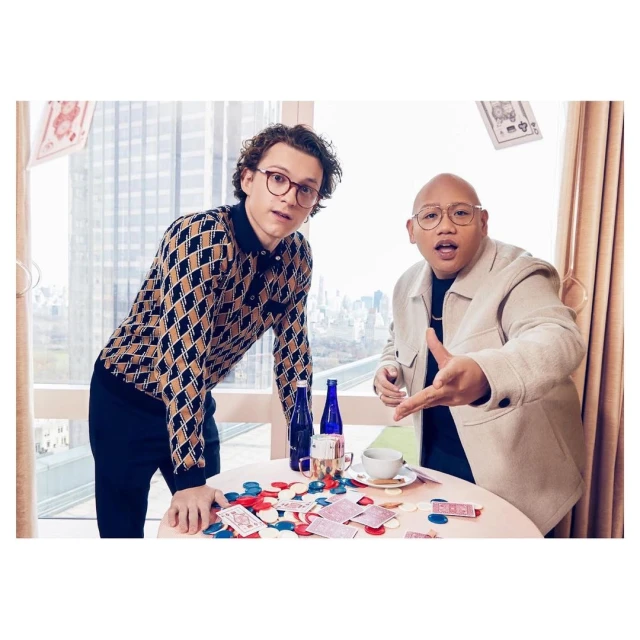Two men, one in patterned polo and the other in a tan jacket, sit at a table with playing cards and chips, with a cityscape background.
