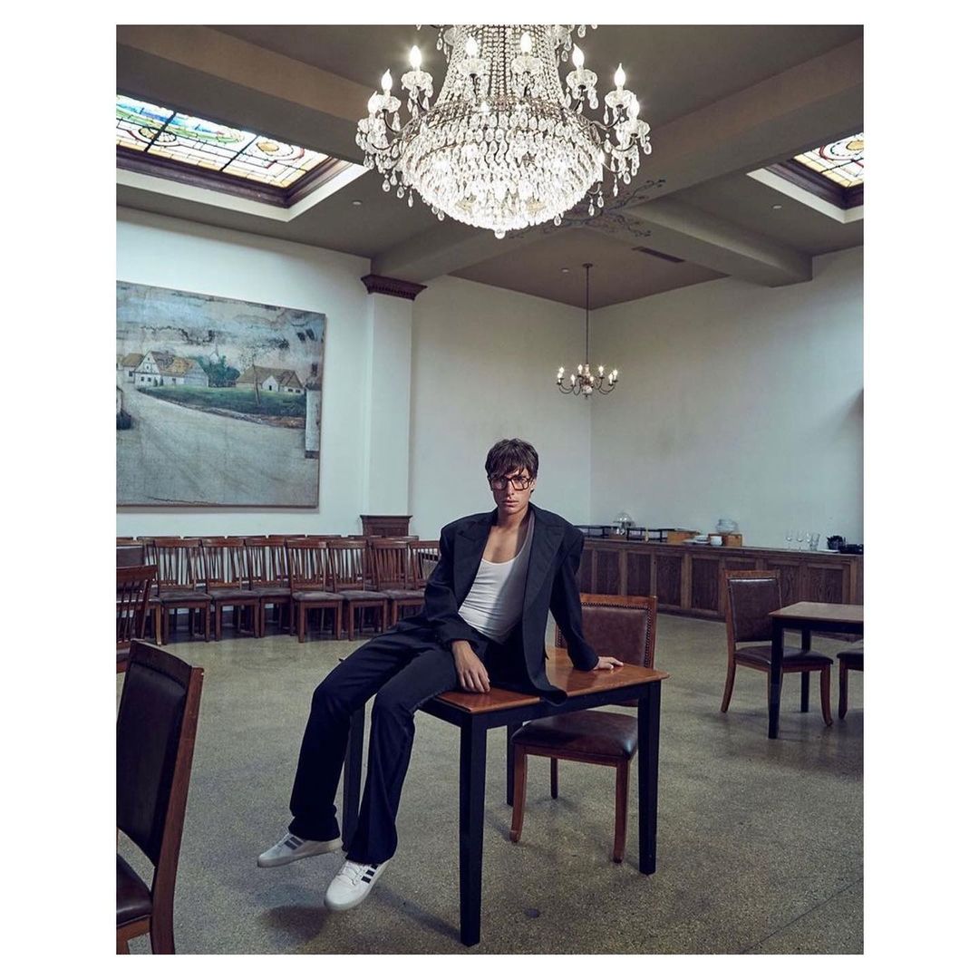 Stylish man in suit posing on table with ornate chandelier, classical interior, and large painting in the background, fashion photography.