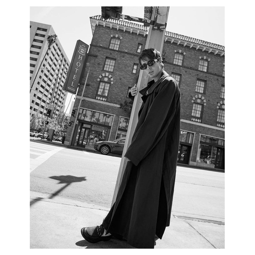 Black and white portrait of a man in sunglasses and a long coat standing on a city street corner with buildings in the background.
