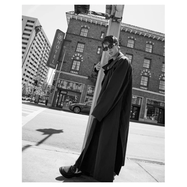 Black and white portrait of a man in sunglasses and a long coat standing on a city street corner with buildings in the background.